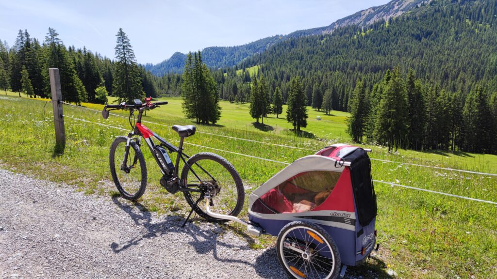 Mountainbike mit Kinderanhänger auf einem Kiesweg im Tiroler Frühling mit grünen Wiesen und Waldlandschaft.